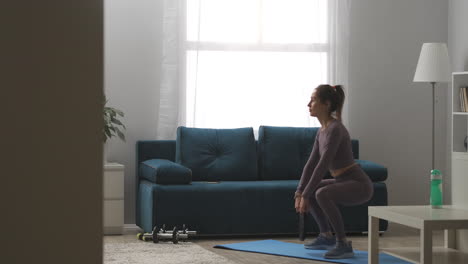 sexy-lady-with-ponytail-is-doing-squats-with-weight-in-hands-training-buttocks-and-legs-muscles-home-fitness-exercises-full-length-shot-in-apartment